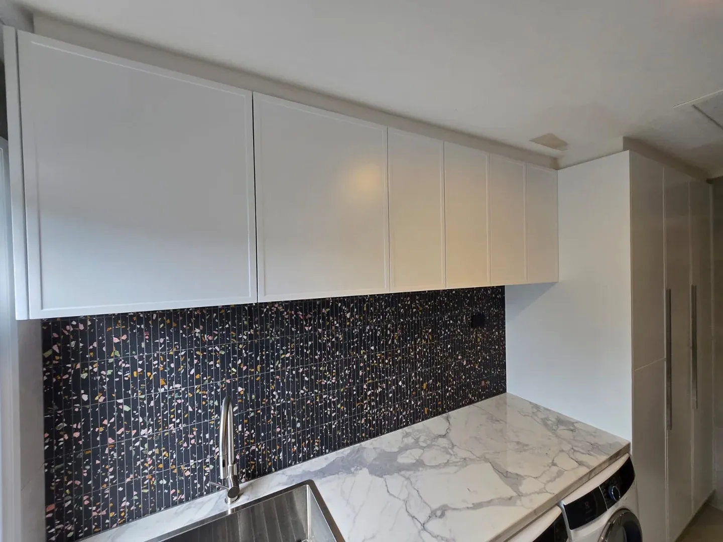 White overhead laundry cupboards above a mosaic splashback and stone benchtop